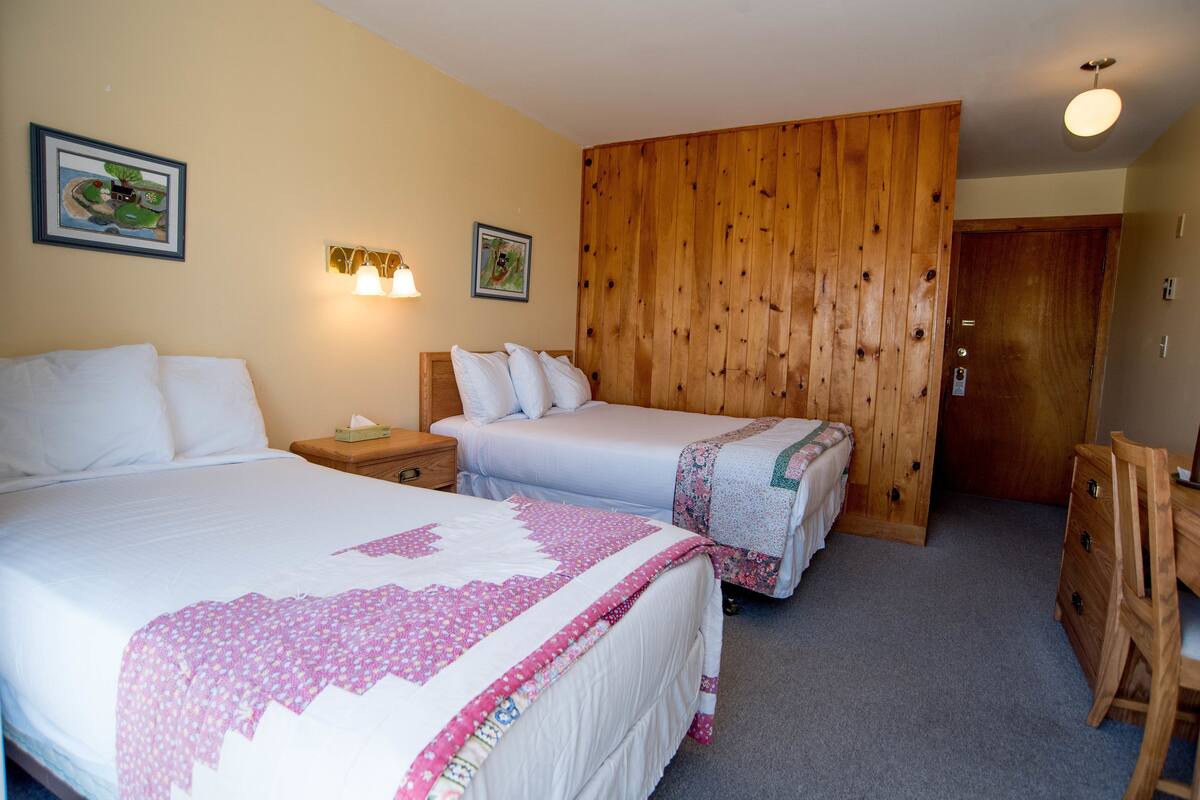 Quarto de hotel com duas camas, lambris de madeira em uma parede e uma escrivaninha com uma cadeira.