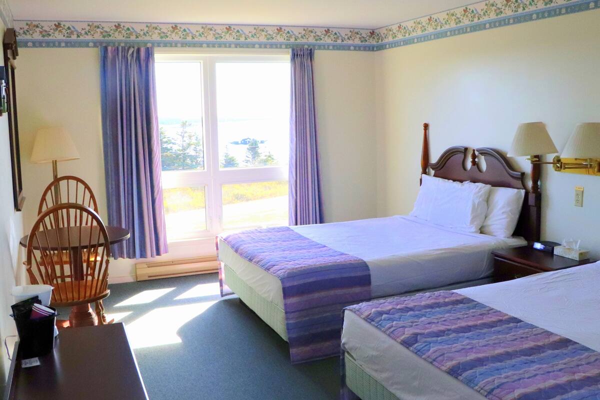 Quarto de hotel com duas camas, uma cadeira, uma mesa e uma janela com cortinas.