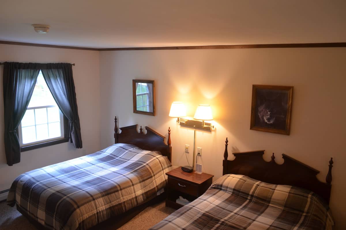 A hotel room with two beds, a nightstand with a lamp, a window with curtains, and a framed picture on the wall.