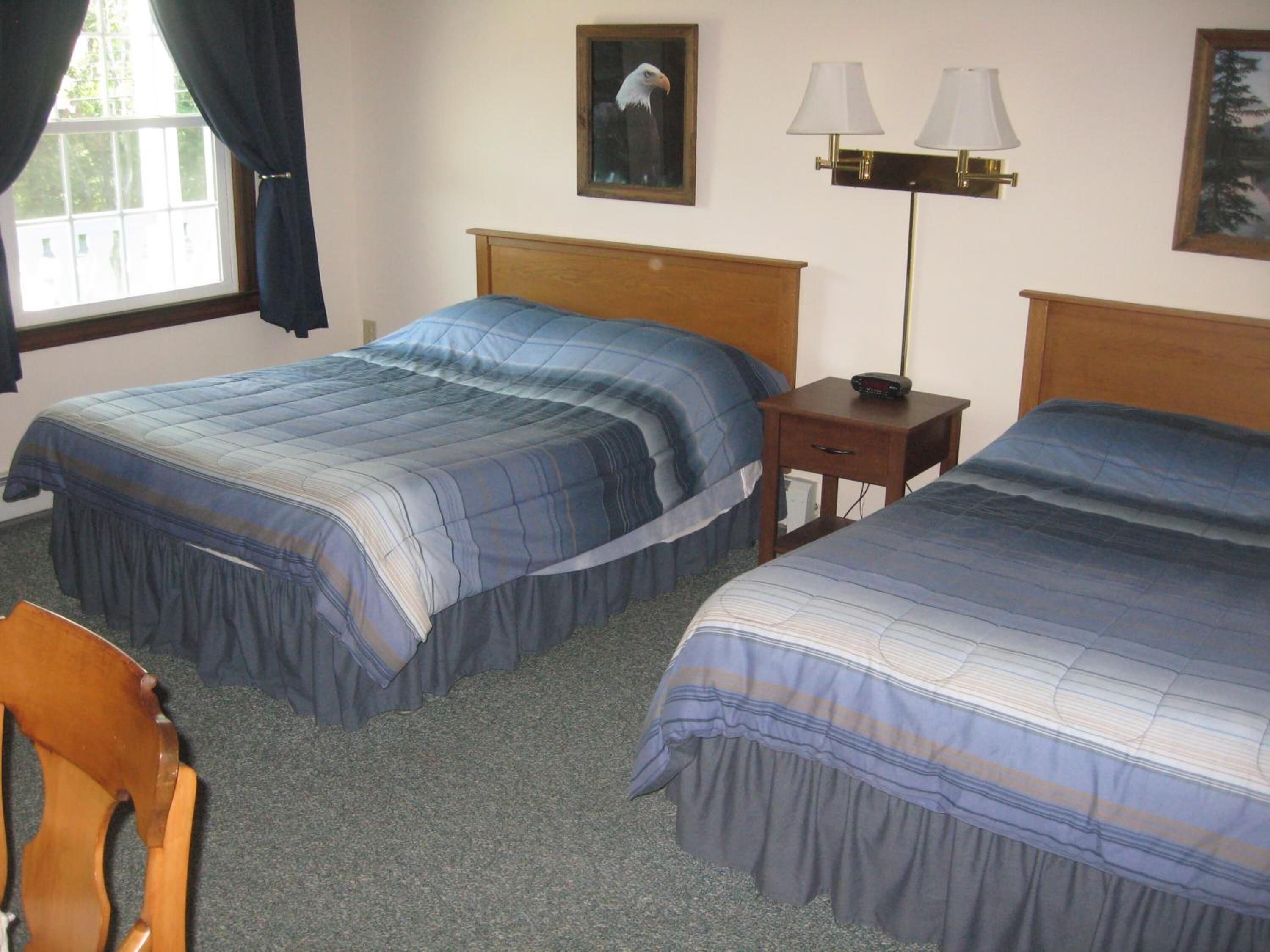Habitación de hotel con dos camas, cabecero de madera, mesita de noche con lámpara y una silla.