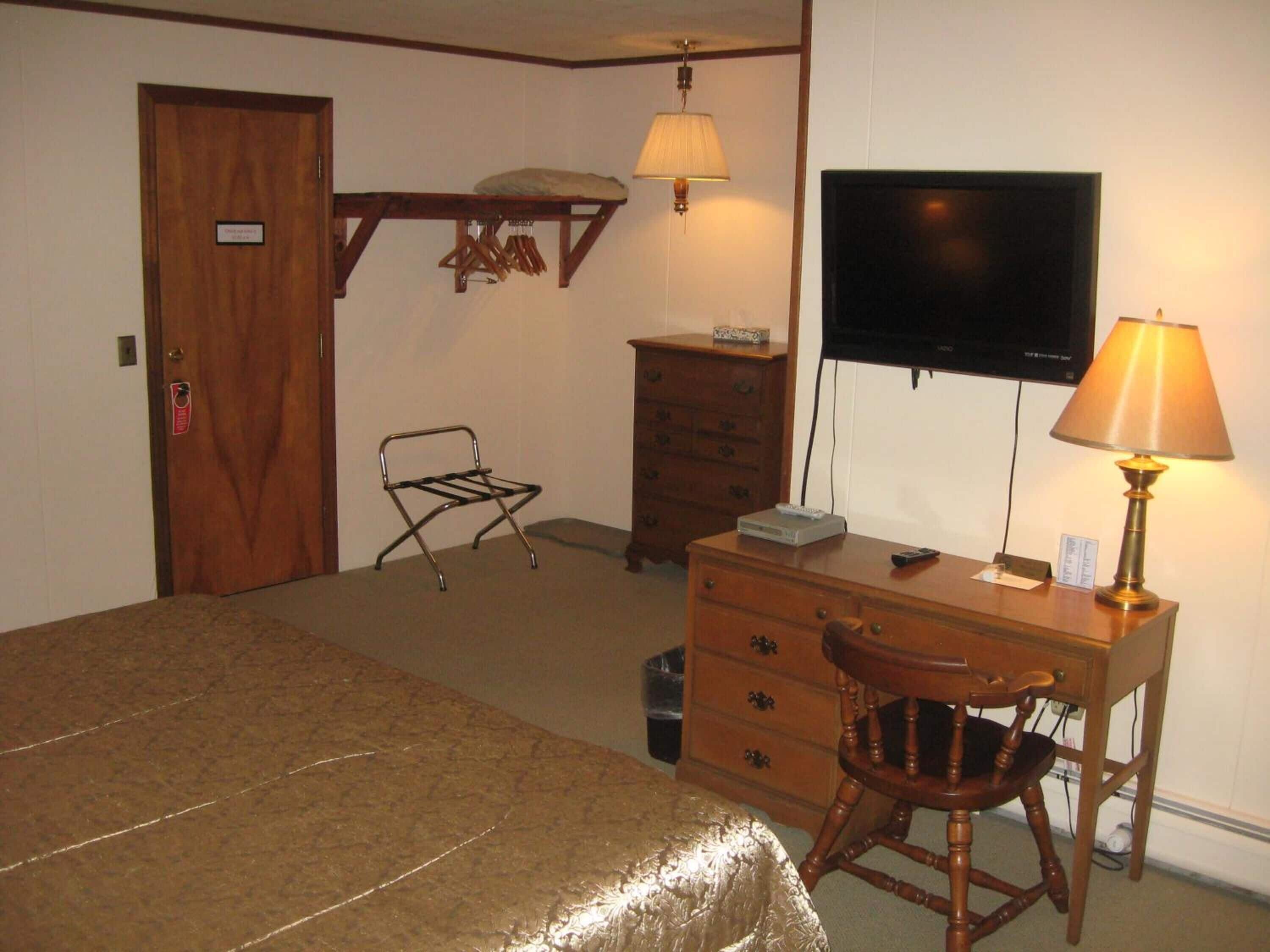 A hotel room with a bed, a wooden desk with a lamp, a television, a wooden chair, and a small table with a remote control.