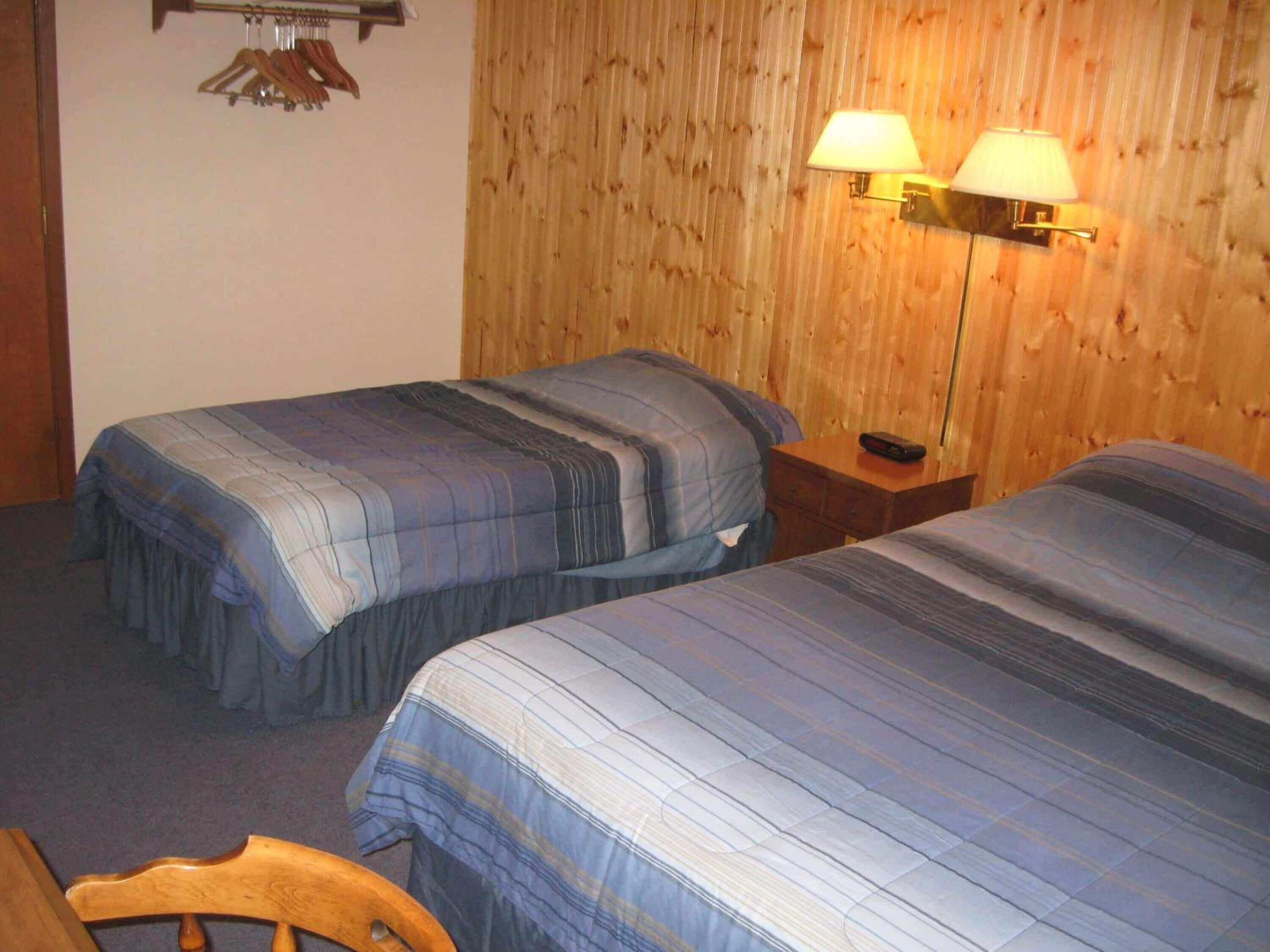 A hotel room with two beds, wooden panel walls, and two wall-mounted lamps.