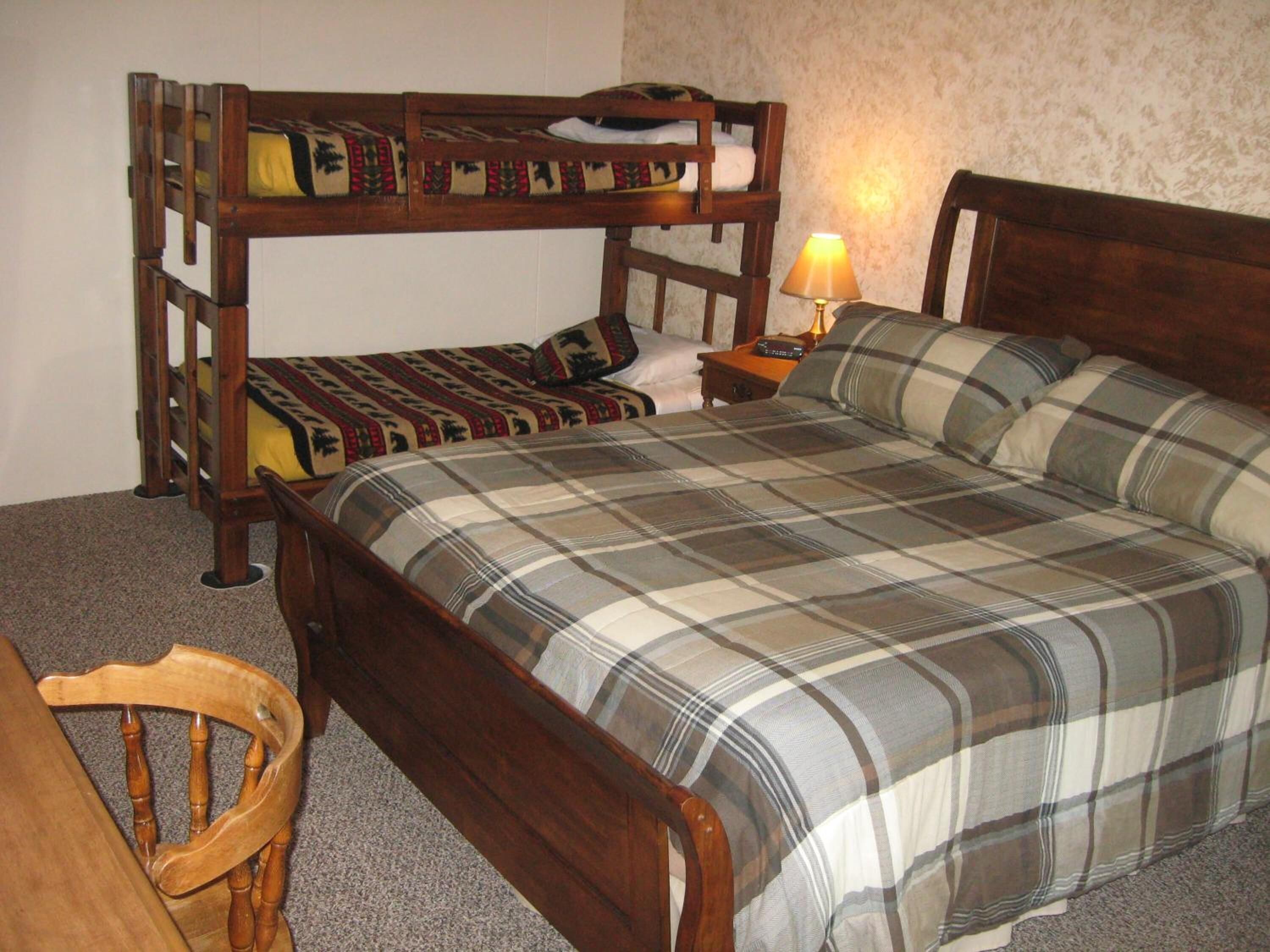 Una cama litera con estructura de madera y una colcha a cuadros. Hay una mesita de noche con una lámpara y una silla.