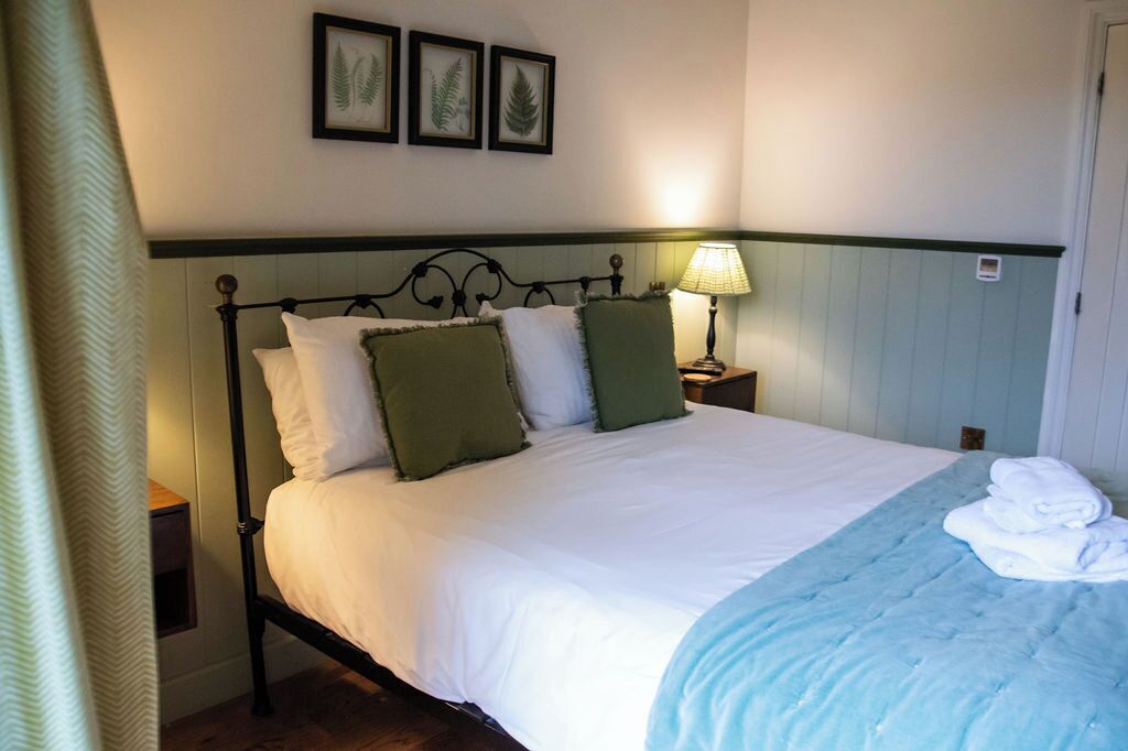 Una cama bien hecha con almohadas blancas y verdes, una manta azulada y una toalla doblada al lado. Una lámpara de noche ilumina un mesita de madera.