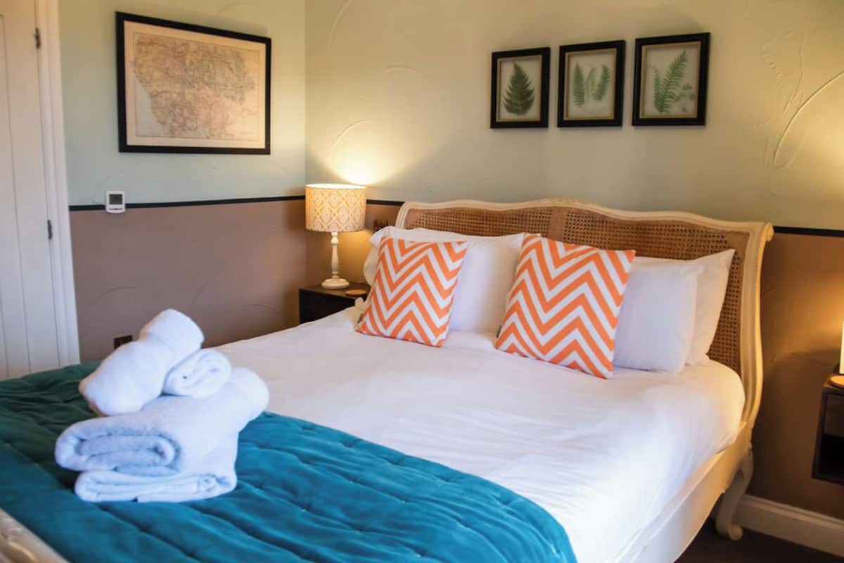 A neatly made bed with white and orange pillows, a teal blanket, and rolled towels on the side.