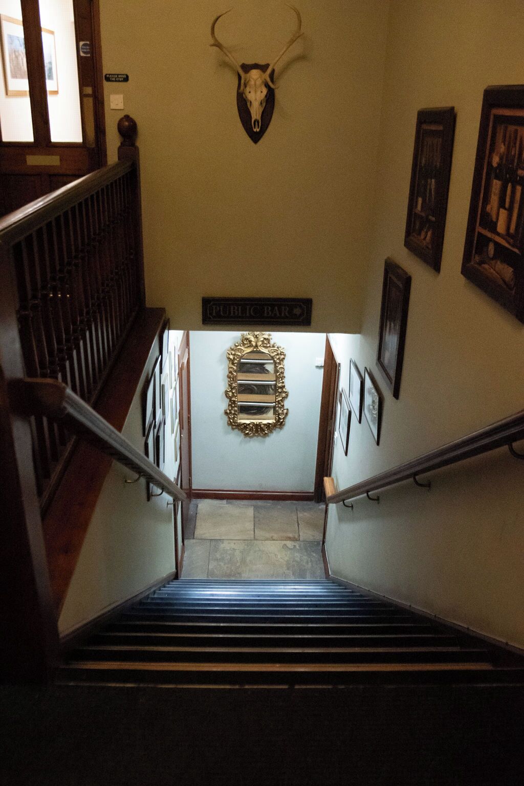 A staircase with a wall-mounted deer skull, a sign reading 'PUBLIC BAR,' and framed artwork on the walls.