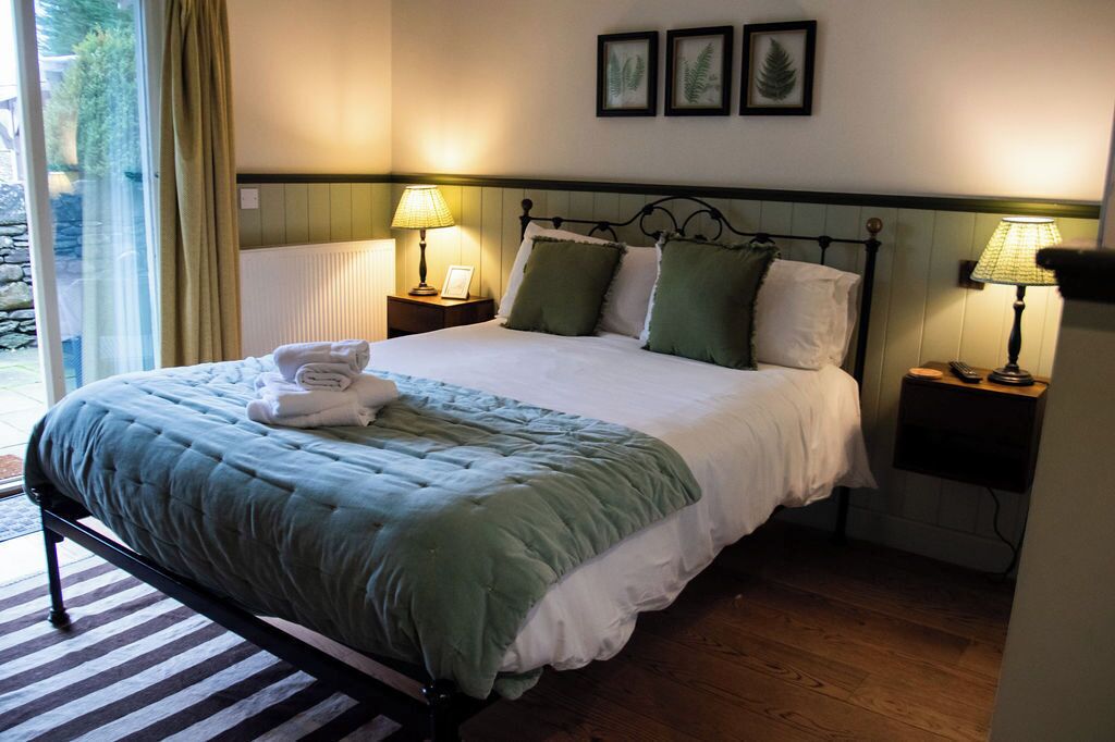 A neatly made bed with a green and white duvet, two bedside lamps, a framed picture on the wall, and a window with curtains.