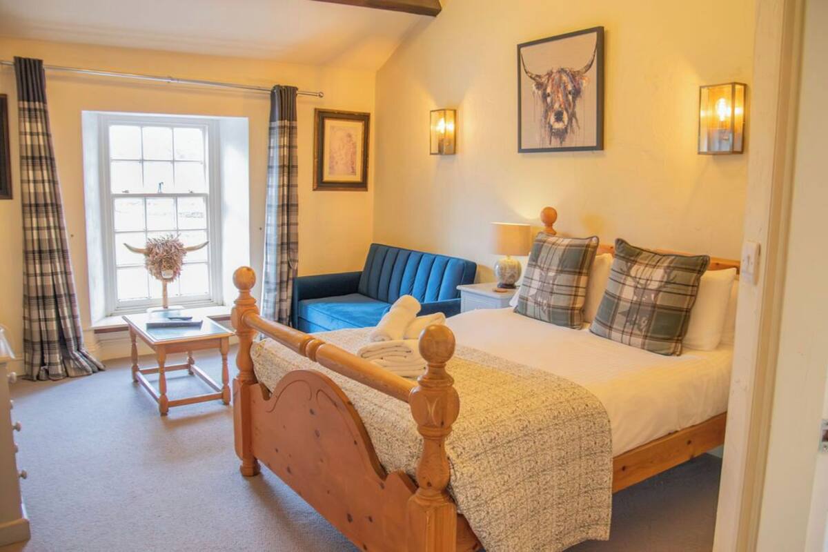 A bedroom with a bed, a blue sofa, a wooden chair, a bedside table with a lamp, and a window with curtains.