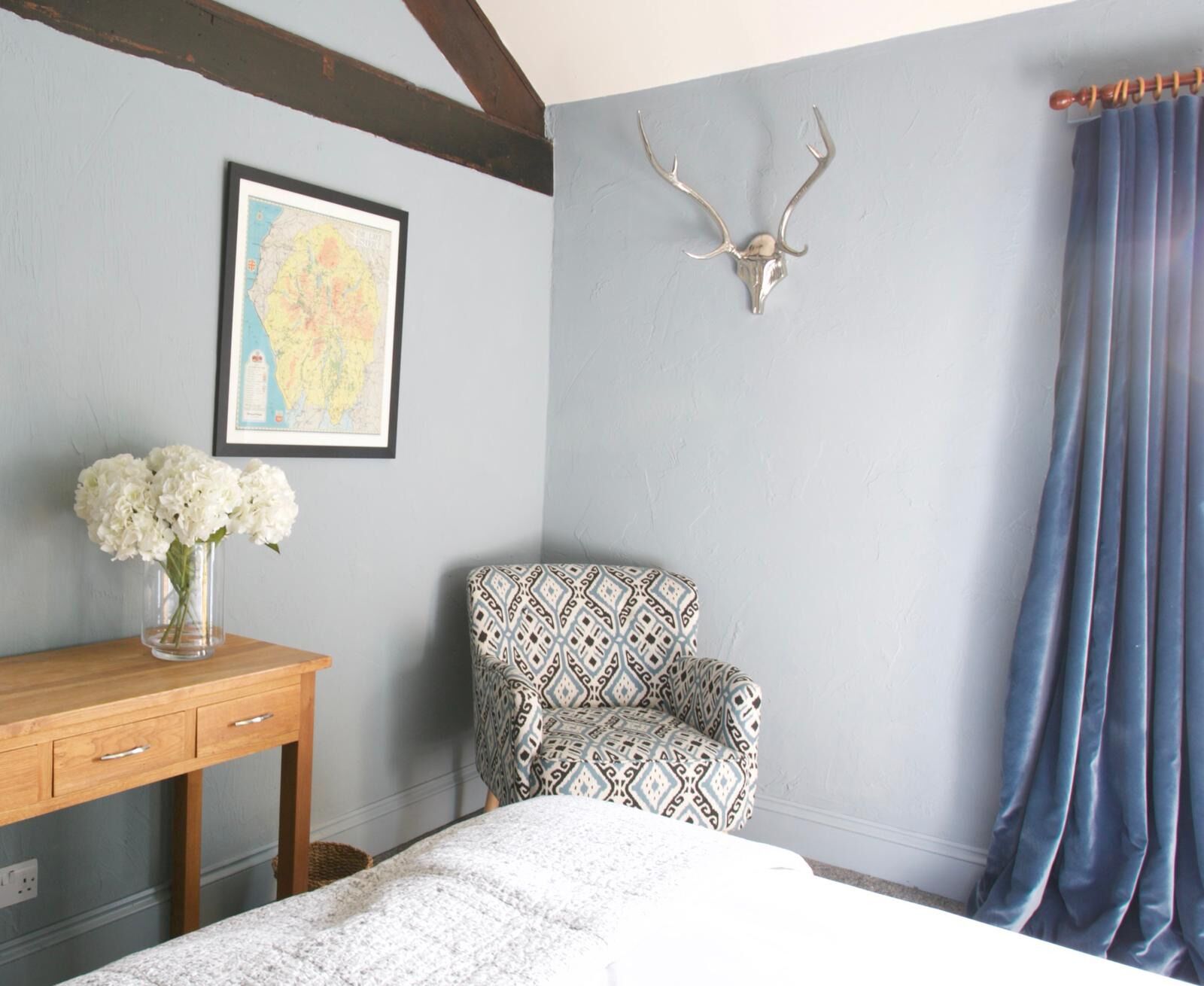 A room with a bed, a desk with a vase of flowers, a patterned armchair, a map on the wall, and a deer skull mounted on the wall.