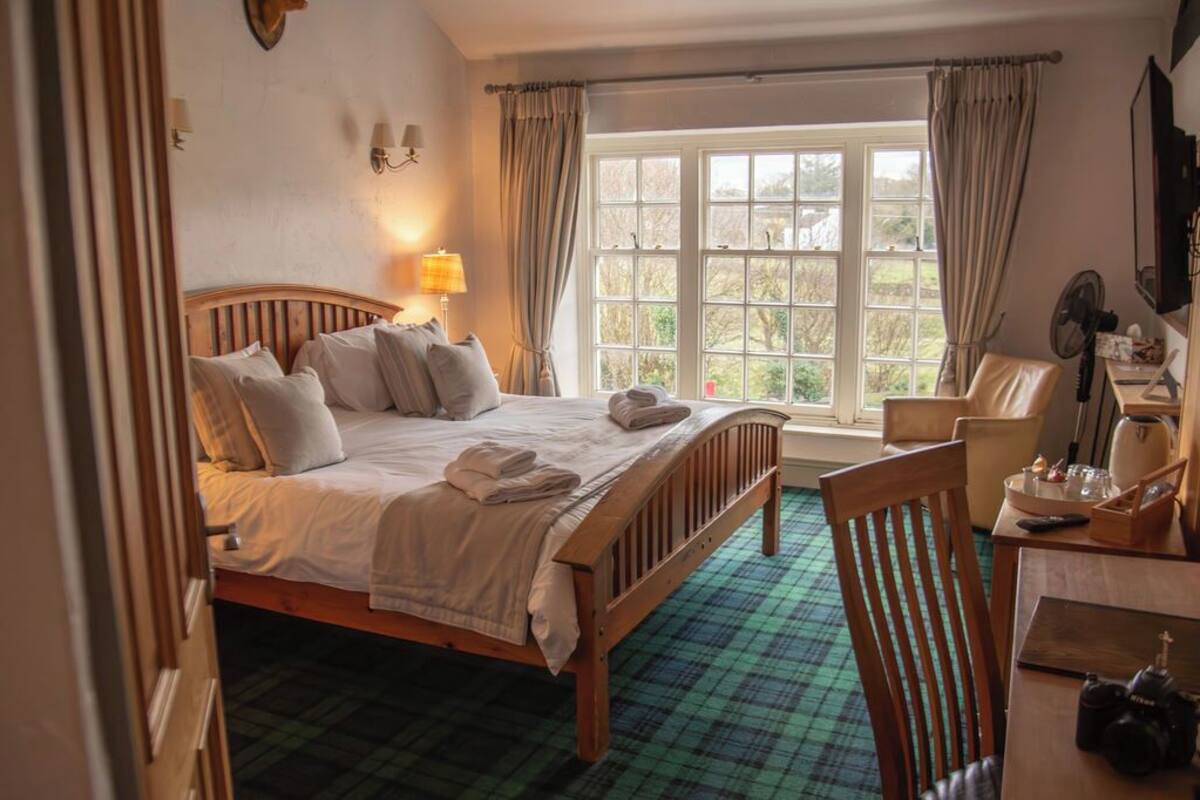 A bedroom with a wooden bed, a window with curtains, a chair, and a table with a fan and a remote control.