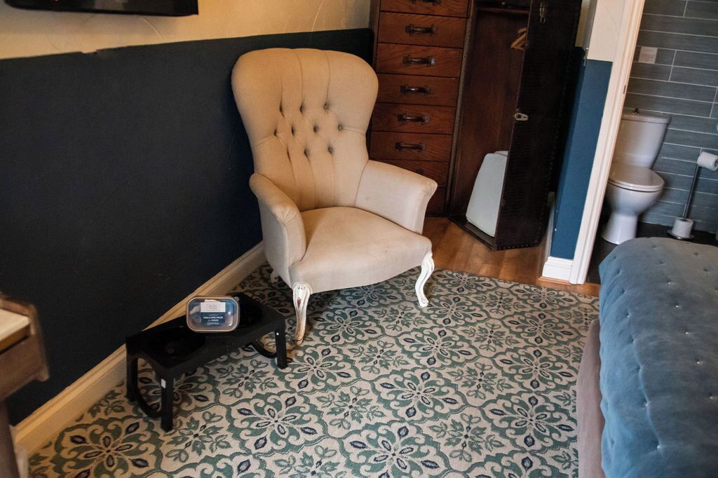 A room with a tufted armchair, a patterned rug, a wooden cabinet, and a blue ottoman.