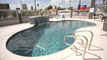 A swimming pool with a small waterfall feature, surrounded by a white fence and a few lounge chairs.