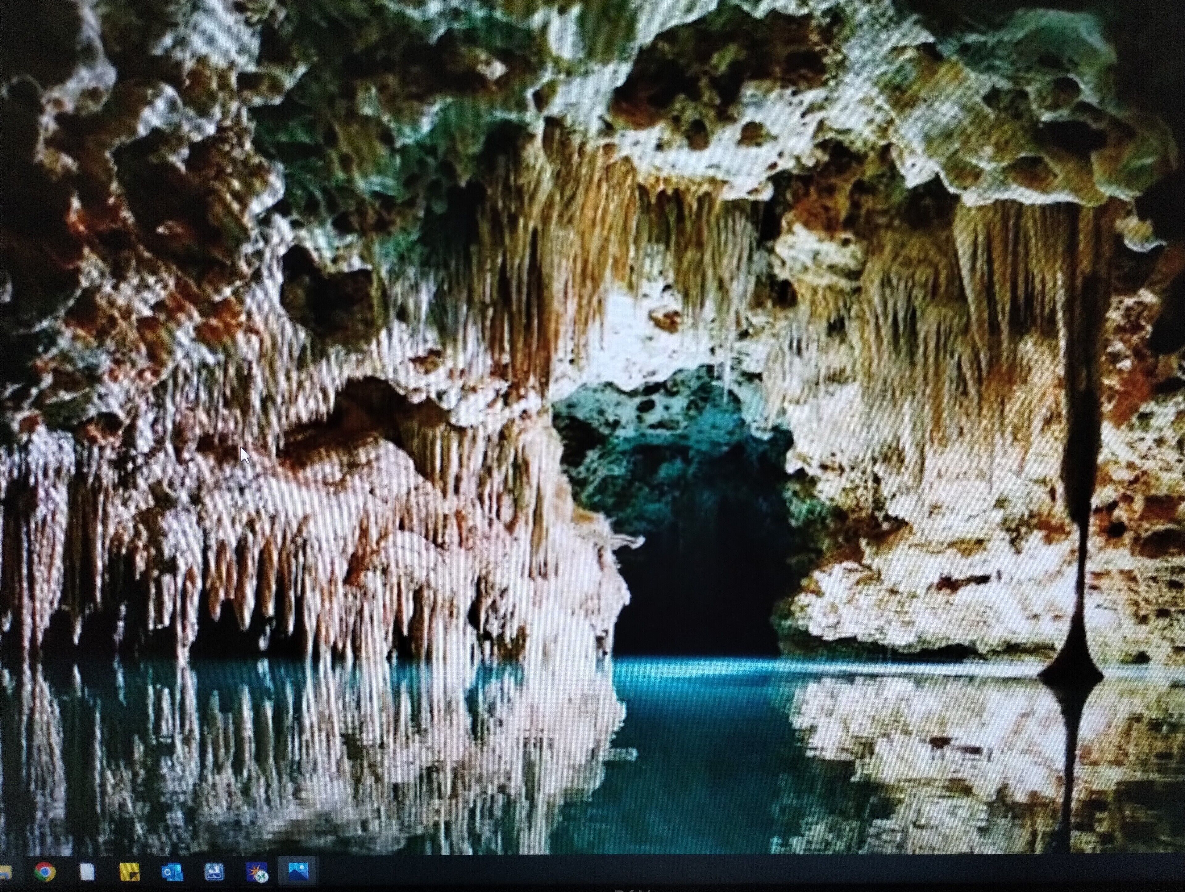 Interior de cueva con estalactitas y estalagmitas, un estanque reflectante y una abertura oscura.