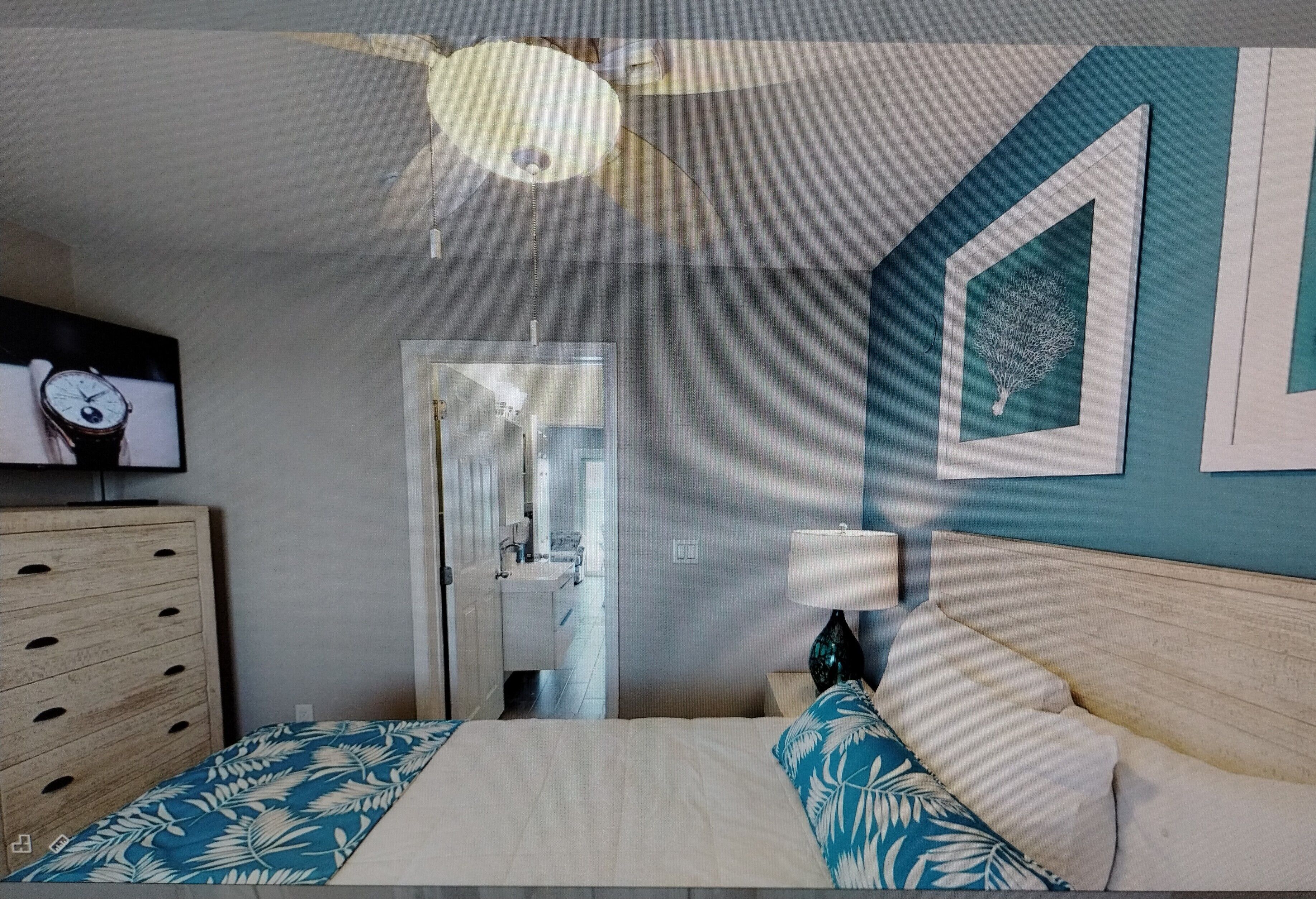 Un dormitorio con cama, cómoda, ventilador de techo, televisor montado en la pared y puerta que da a un baño.