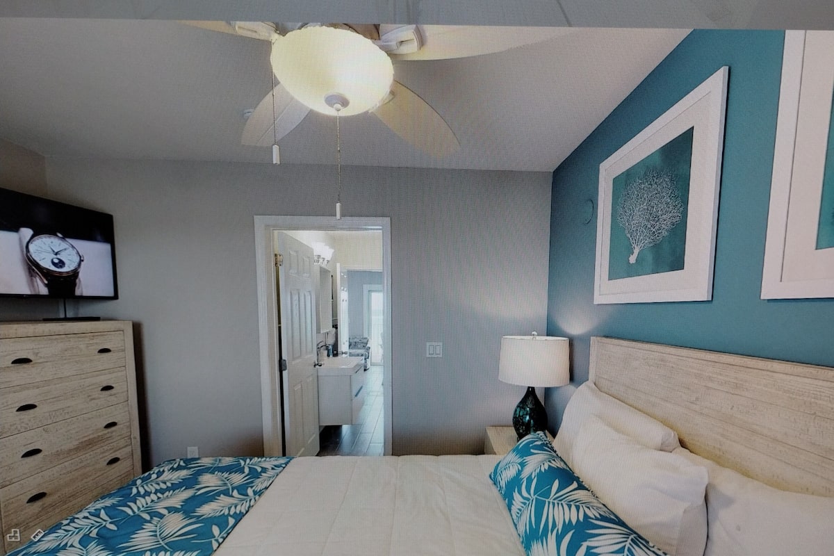 A bedroom with a bed, a dresser, a ceiling fan, a wall-mounted TV, and a door leading to a bathroom.