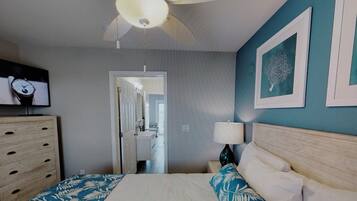 Un dormitorio con cama, cómoda, ventilador de techo, televisor montado en la pared y puerta que da a un baño.