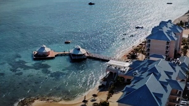 An aerial view of a coastal hotel with multiple domed structures extending into the water, a pier connecting to the beach, and a row of buildings with blue roofs. - Morritts Tortuga Club (East End)
