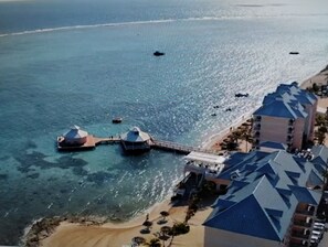 An aerial view of a coastal hotel with multiple domed structures extending into the water, a pier connecting to the beach, and a row of buildings with blue roofs. - Morritts Tortuga Club (East End)