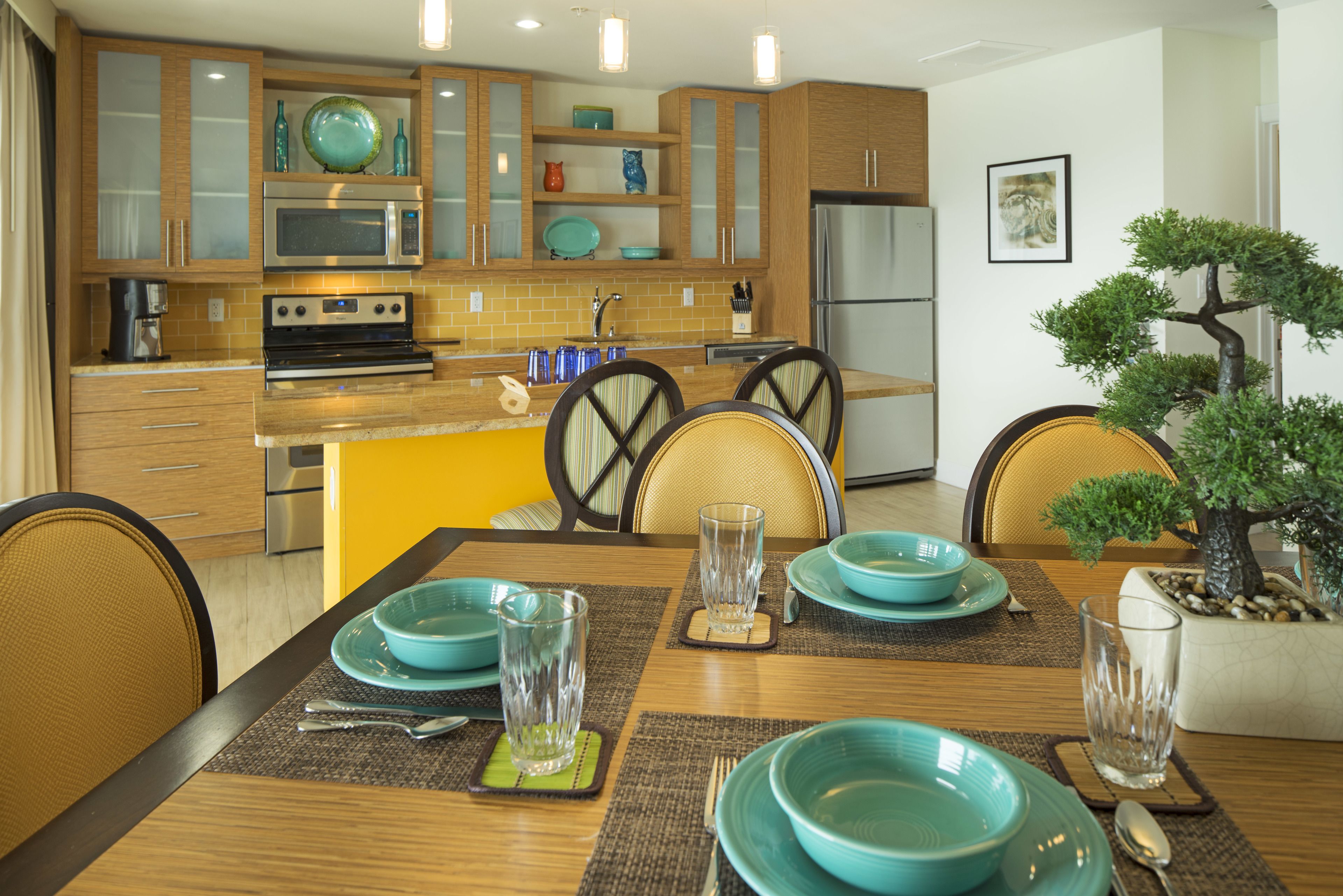 A modern kitchen with wooden cabinets, a stainless steel refrigerator, and a dining table set with blue plates and clear glasses.