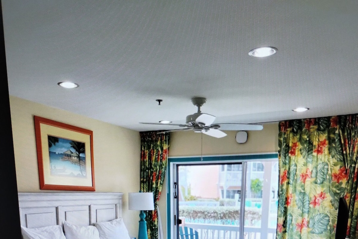 A hotel room with a bed, a nightstand, a ceiling fan, and a view of the outside.