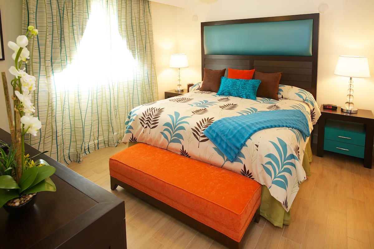 A bedroom with a bed, bedside tables, a nightstand, a bench, a potted plant, and a window with curtains.