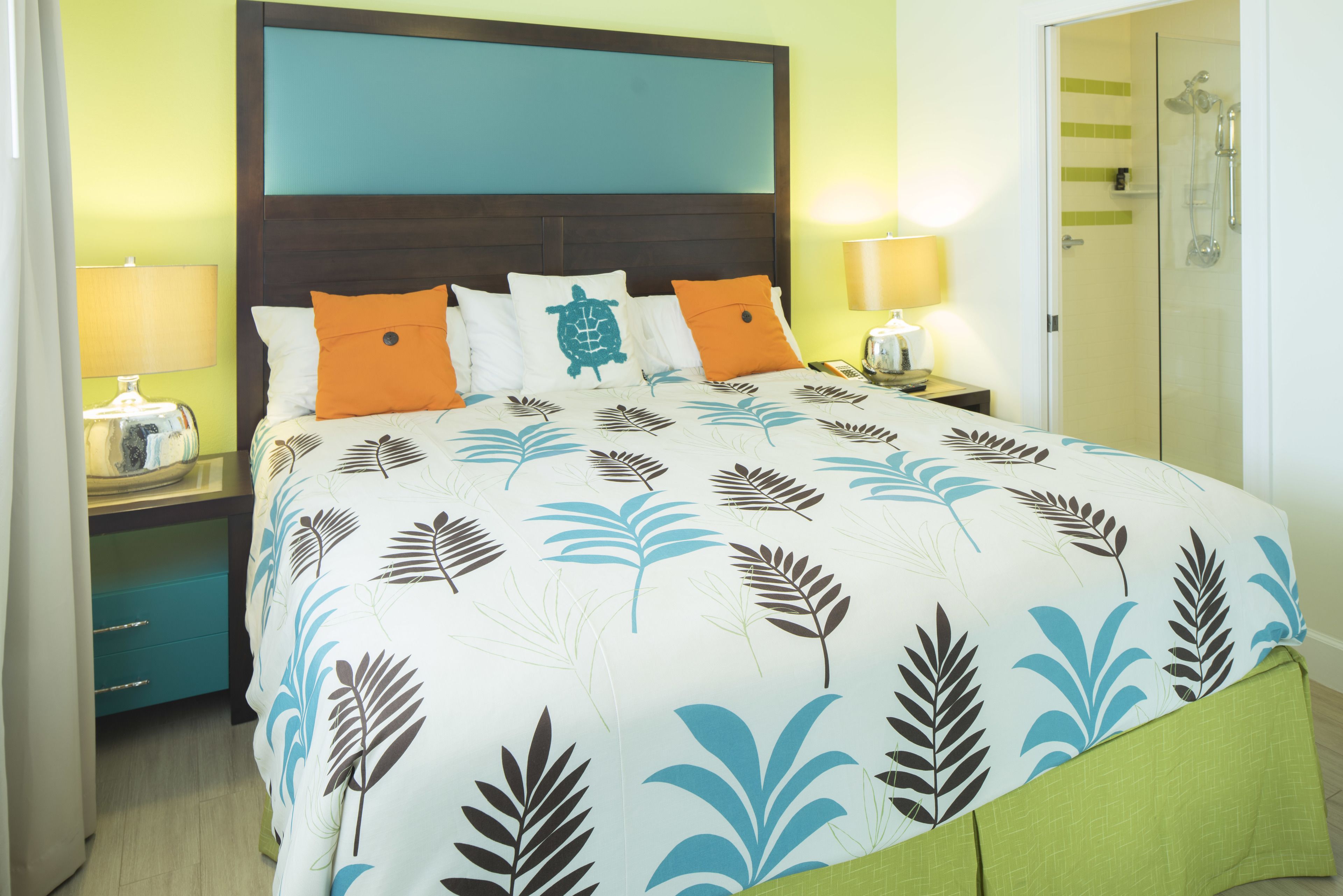 A neatly made bed with a leaf-patterned bedspread, two bedside lamps, a nightstand, and a green drawer unit.