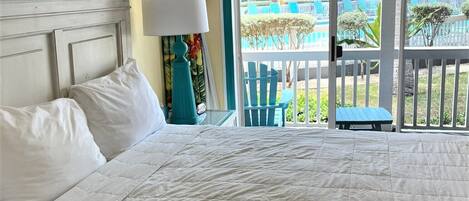 Habitación de hotel con cama, mesita de noche con lámpara y vistas a una piscina y zona de estar exterior.