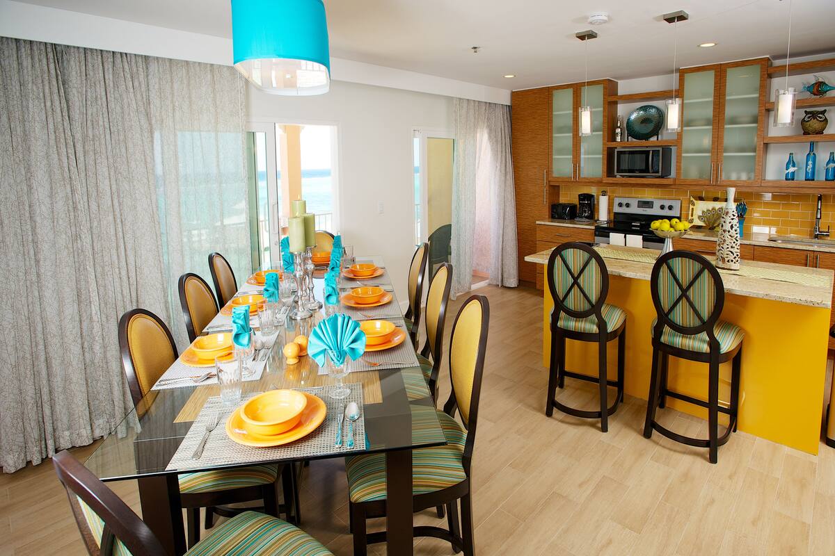 A modern dining area with a glass table, striped chairs, and a kitchen in the background.