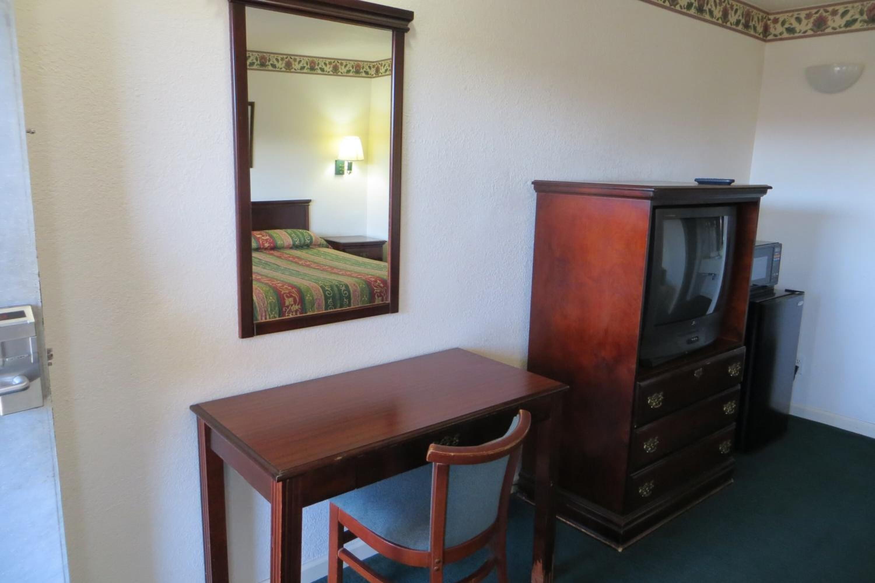 A hotel room with a wooden desk, a chair, a television on a wooden cabinet, and a mirror on the wall.