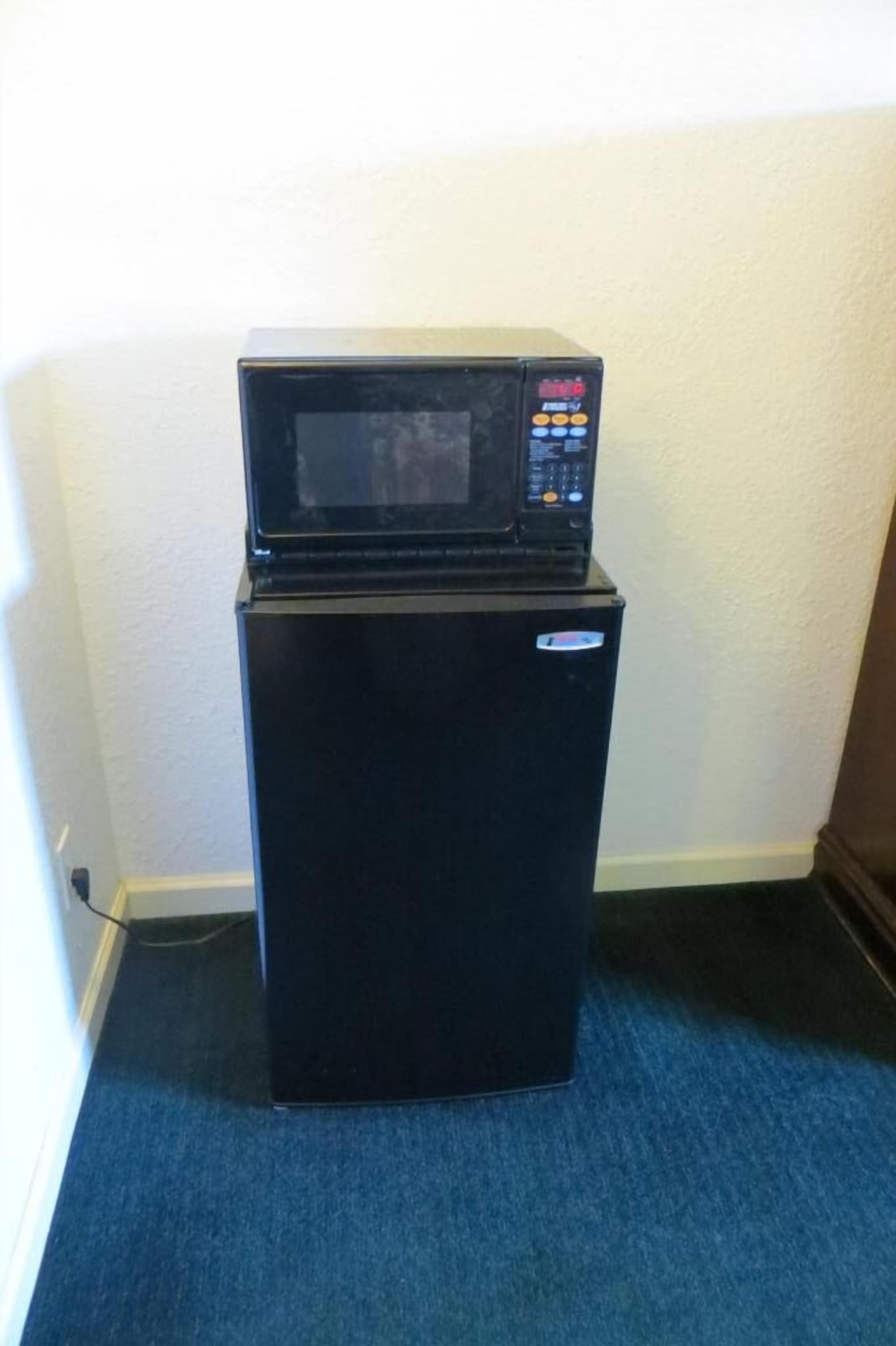 A black refrigerator with a microwave on top, located in a corner with a blue carpet.
