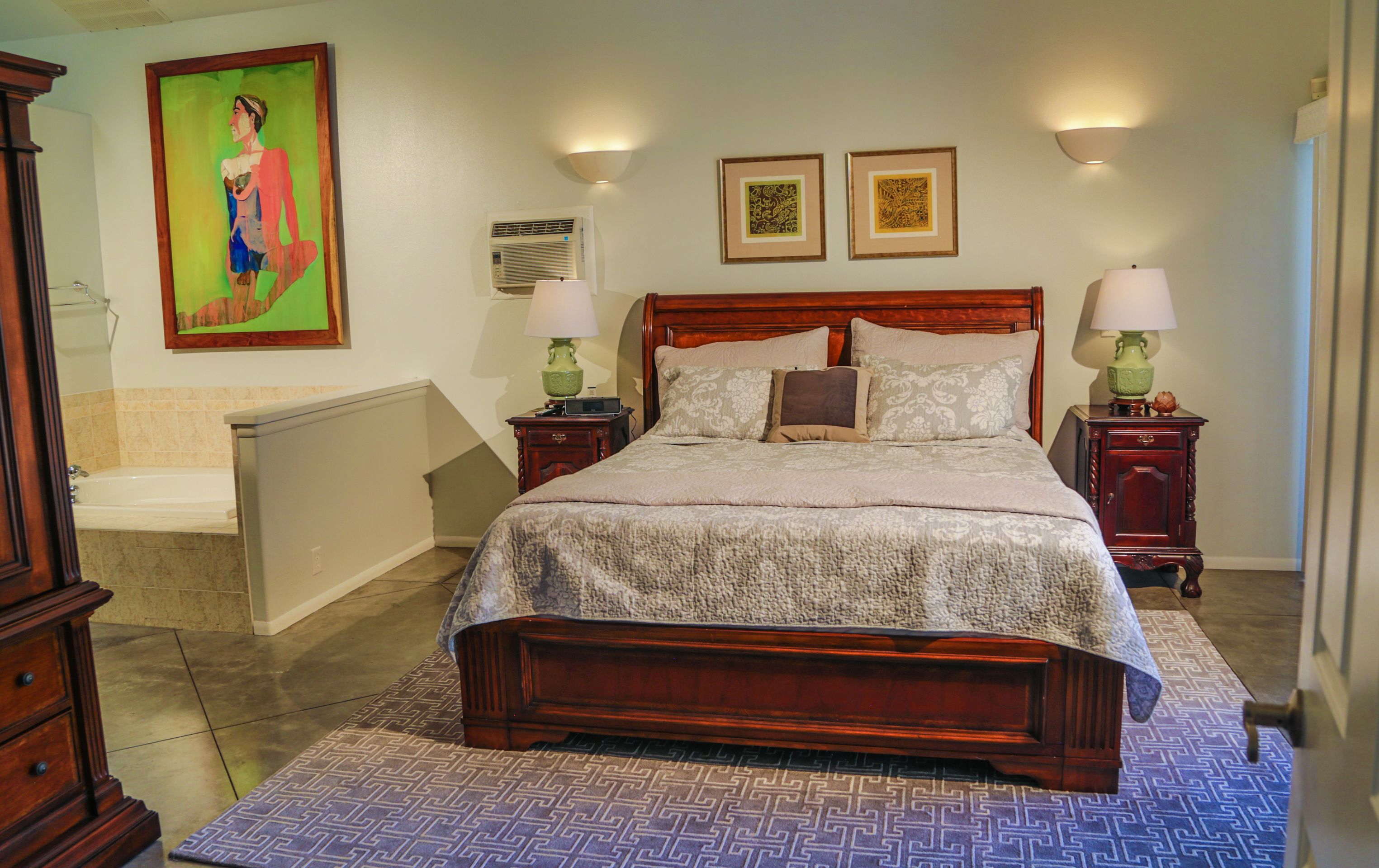 A neatly arranged bedroom with a wooden bed, two bedside tables with lamps, a large painting on the wall, and a bathroom with a bathtub and shower.