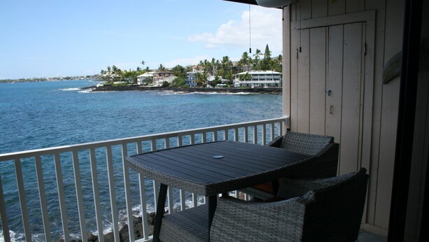 A balcony with a table and chairs overlooking a coastal area with houses and the sea. - Sea Ge 1-203 1 Bedroom Condo by RedAwning (Kailua-Kona)