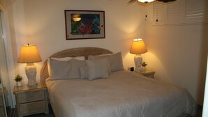 A neatly made bed with a headboard, two bedside tables with lamps, a framed picture on the wall, and a window with blinds. - Sea Ge 1-203 1 Bedroom Condo by RedAwning (Kailua-Kona)