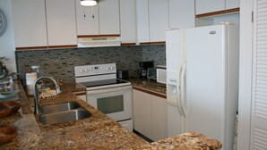 A kitchen with granite countertops, white cabinets, a built-in oven, a refrigerator, and a coffee maker. - Sea Ge 1-203 1 Bedroom Condo by RedAwning (Kailua-Kona)