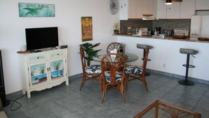A living room with a TV, a dining area with chairs, a kitchen with a counter, and a ceiling fan. - Sea Ge 1-203 1 Bedroom Condo by RedAwning (Kailua-Kona)
