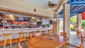 A bar area with wooden stools and a round table, surrounded by stone flooring and a covered patio with chairs.