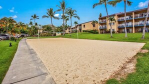 A sandy beach volleyball court with a net, surrounded by palm trees and a hotel building with balconies.