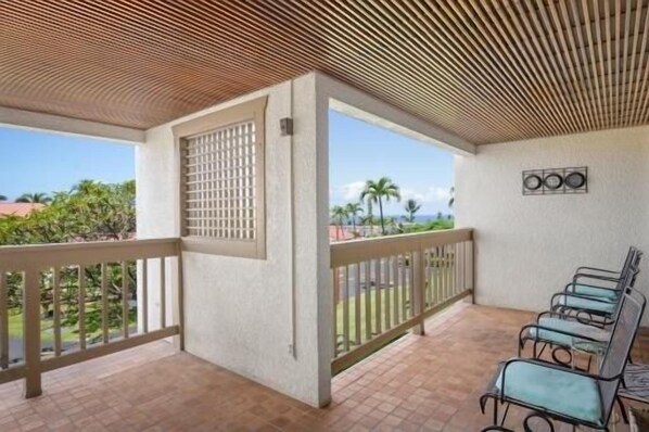 A covered outdoor area with a wooden ceiling, a seating area with chairs, and a view of palm trees and the ocean.