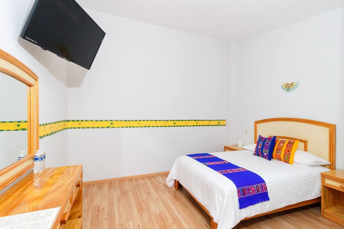 A hotel room with a bed, wooden headboard, bedside tables, a mirror, a flat-screen TV mounted on the wall, and a decorative strip on the wall.