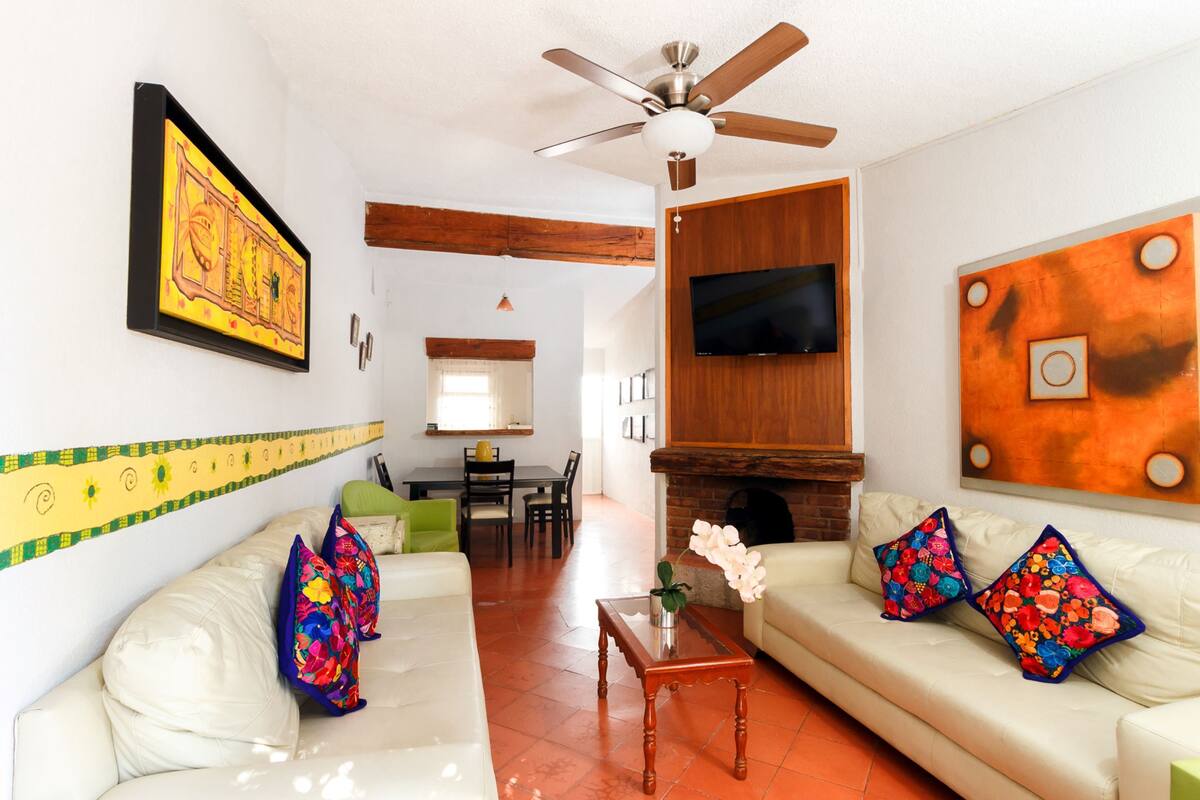 A living room with a sofa, coffee table, and decorative pillows. There is a television mounted on the wall and a ceiling fan.