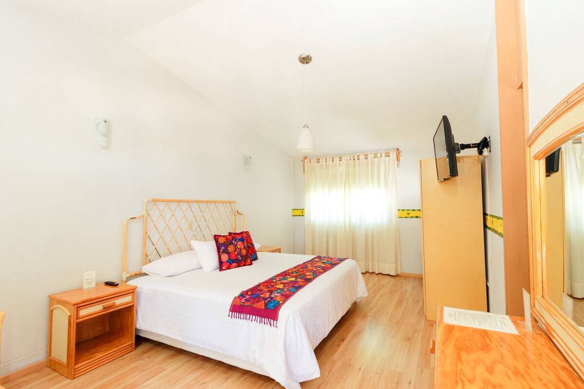 A bedroom with a wooden bed frame, white bedding, a colorful throw blanket, a nightstand, a mirror, and a window with curtains.