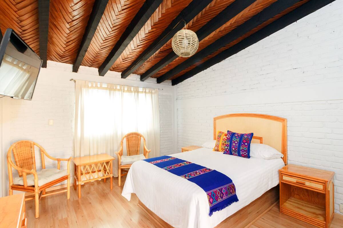 A bedroom with a wooden ceiling, a bed with a colorful blanket, two wicker chairs, a small table, and a nightstand.