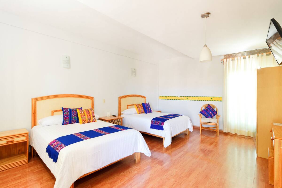 A hotel room with two single beds, wooden floors, a wooden nightstand, a chair, and a TV mounted on the wall.