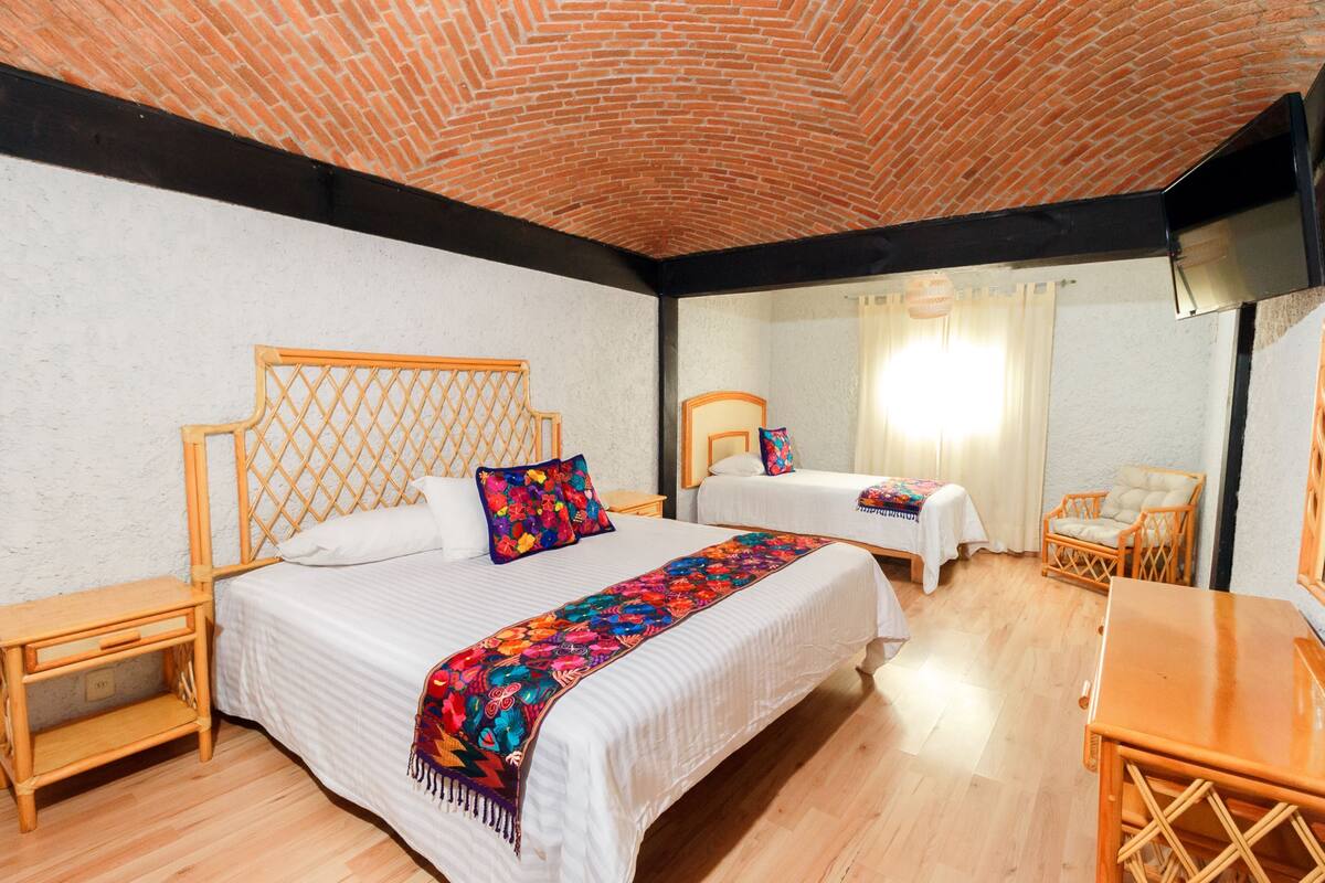 A hotel room with two beds, wooden furniture, a brick ceiling, and a large window.