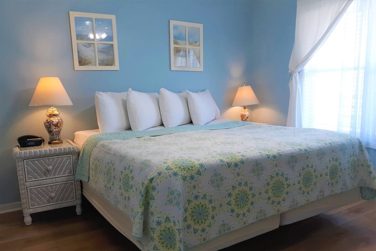 Une chambre à coucher avec un lit, deux tables de chevet, deux lampes, une horloge et deux tableaux encadrés accrochés au mur.