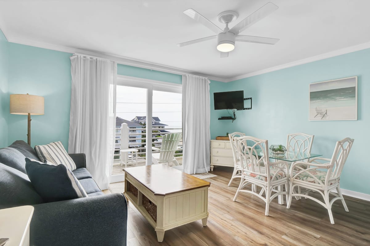Un salon comprenant un canapé, une table basse avec un plateau en verre et un coin repas avec des chaises en osier blanc et une table en verre.