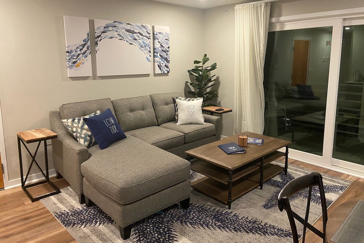 Un salon moderne avec un canapé d’angle gris, une table basse en bois et une petite table d’appoint. On y trouve des œuvres d’art abstraites accrochées au mur et une plante en pot.