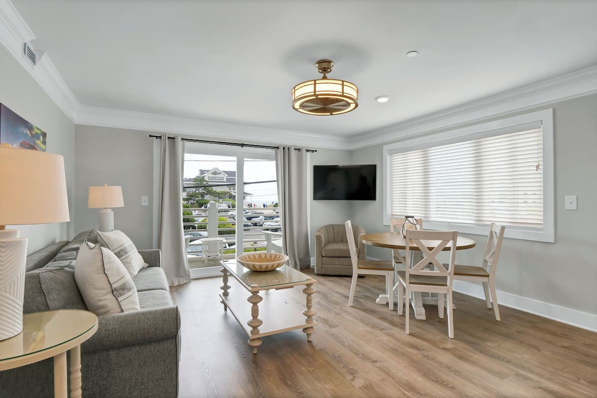 Un salon moderne avec un canapé gris, une table basse en verre, une table à manger avec des chaises, un téléviseur fixé au mur et une grande fenêtre avec des stores.
