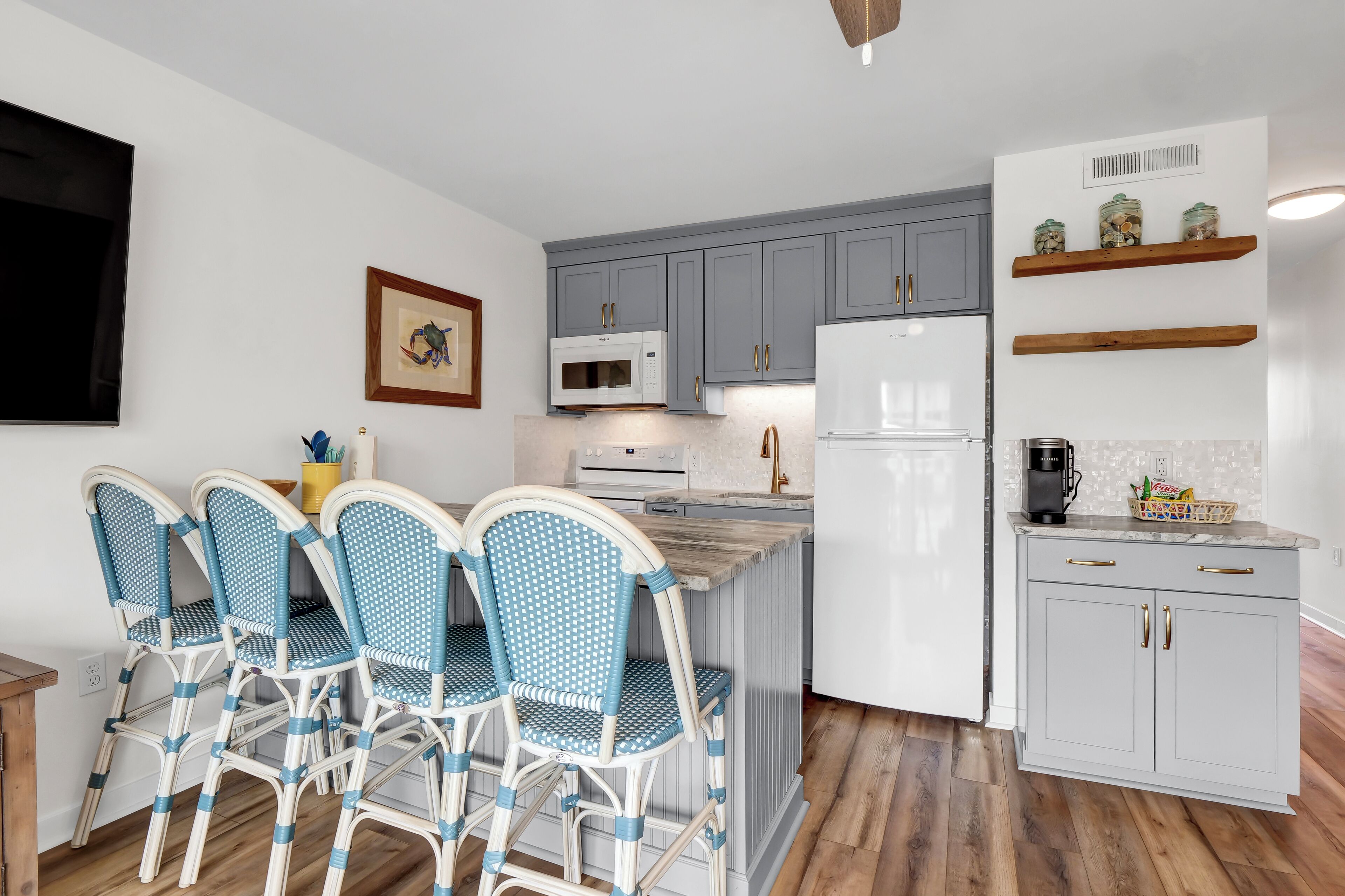 Une cuisine avec des meubles gris, un micro-ondes blanc, un réfrigérateur blanc et un plan de travail en bois. On y trouve deux chaises en osier avec des coussins bleus.