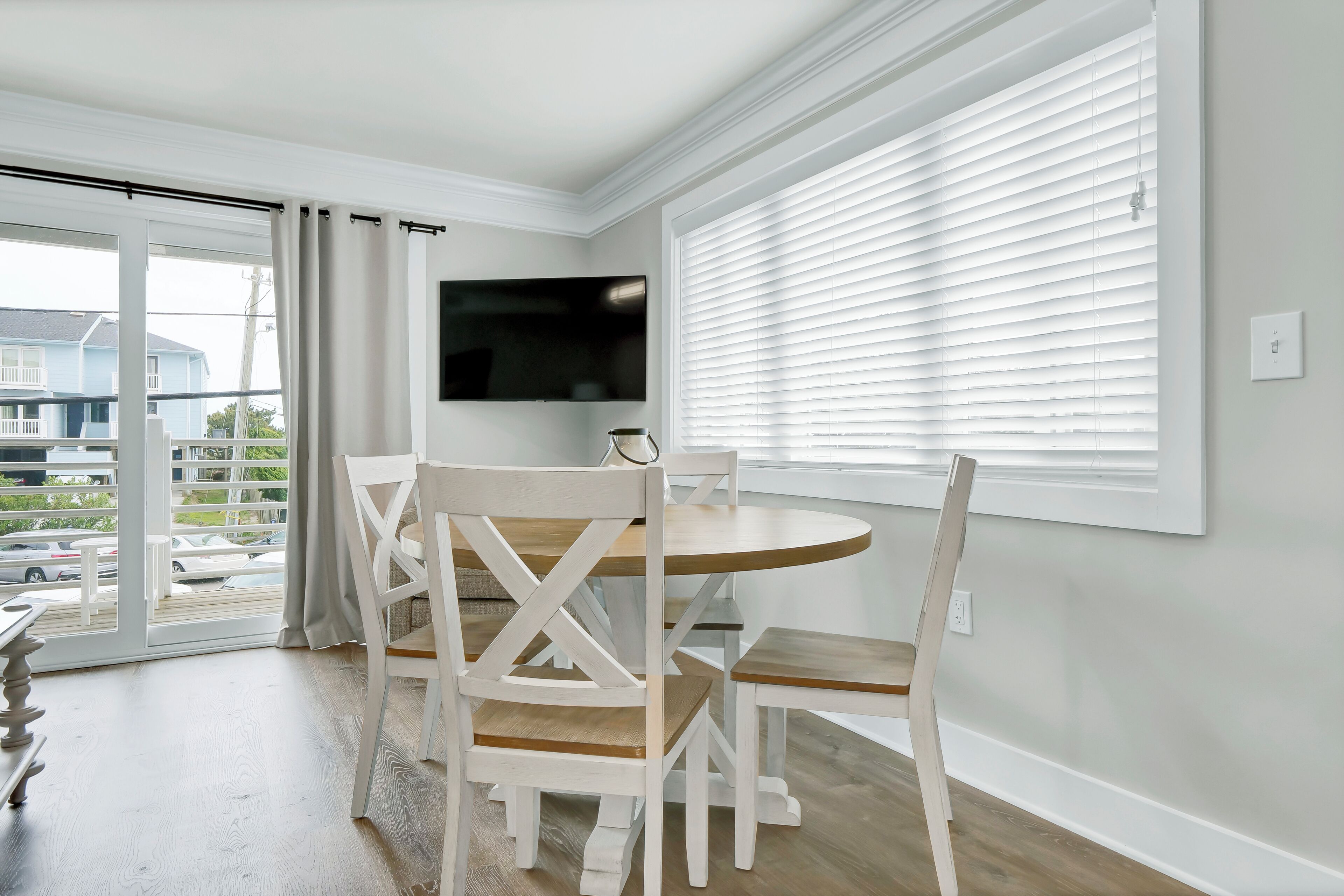 Un coin repas avec une table ronde et des chaises blanches, un téléviseur fixé au mur et une vue sur l’extérieur grâce à une porte-fenêtre coulissante.